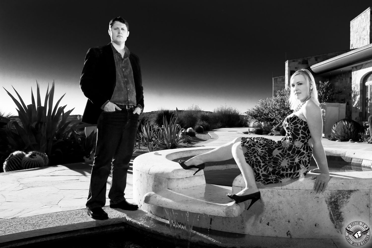 Seductive blond girl in flower print knee length black and white dress with black high heels sits on the edge of a stone fountain near a stoic guy with dark hair wearing a dark sport coat over a grey shirt with dark slacks on a stone poolside walkway in front of cactus and a big dark sky in this engagement photo in Dripping Springs Texas.  